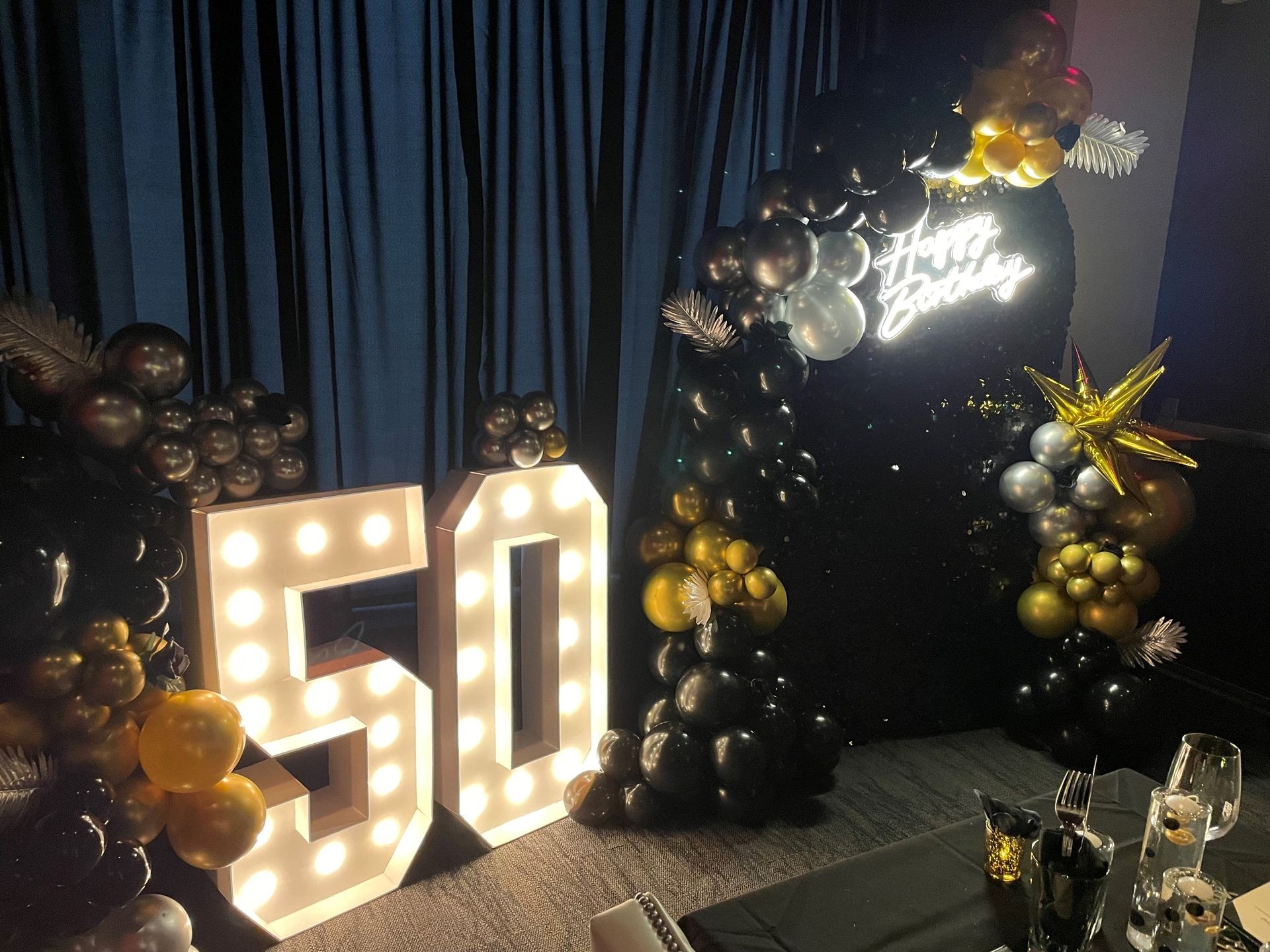 Birthday celebration setup with illuminated number 50, balloon decorations, and a neon Happy Birthday sign.